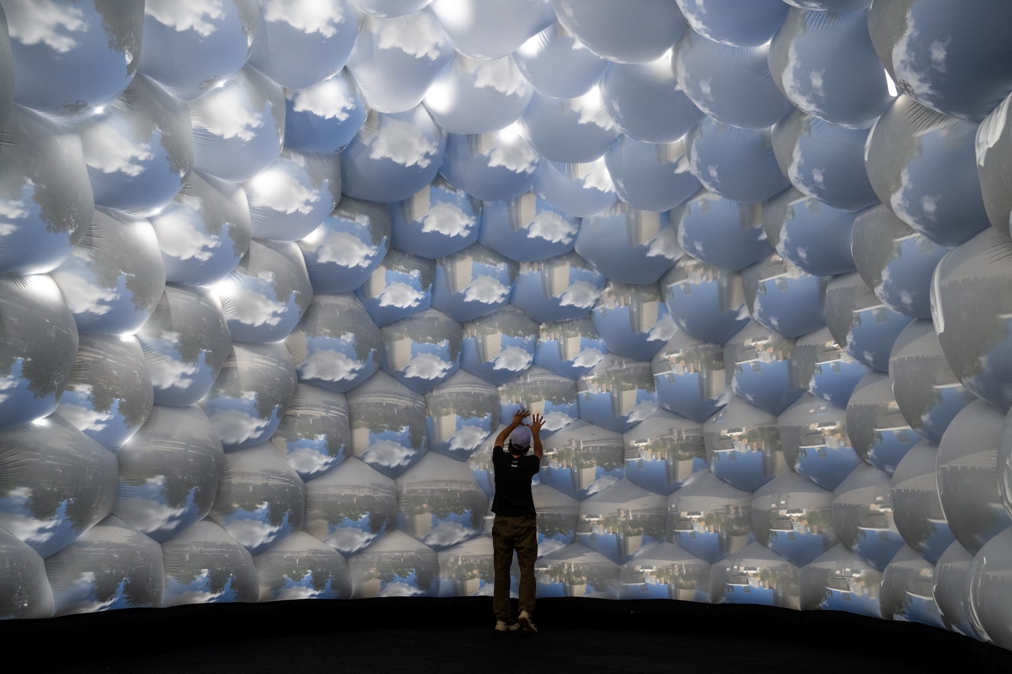 A visitor stands inside Compound Camera No.3 beneath rounded surfaces carrying repeated projections of clouds and buildings.