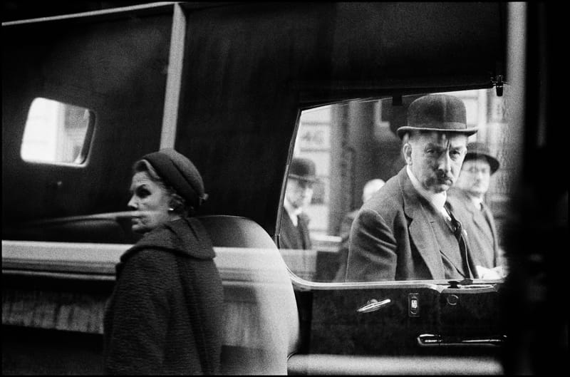 A man and woman appear in layered reflections across the window and body of a black car in London