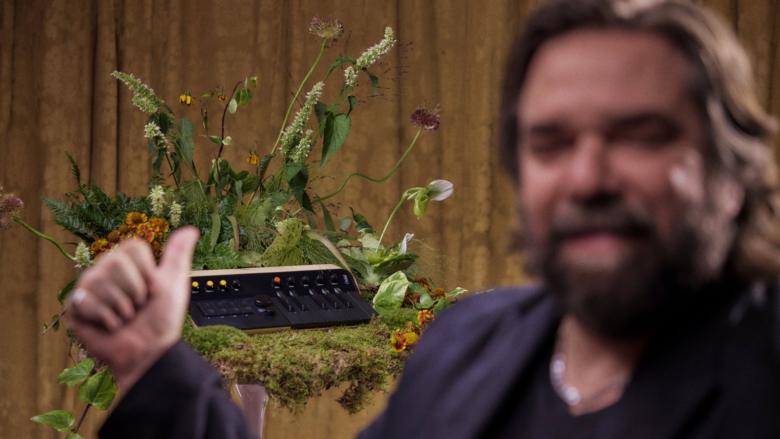 Kevin Parker gesturing toward an Orchid synthesizer arranged among flowers and moss